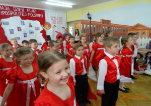 Na tle dekoracji z mapą Polski i dziećmi z chorągiewkami przedszkolaki stoją w rzędach.